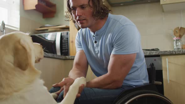 Happy caucasian disabled man in wheelchair playing with pet dog in kitchen