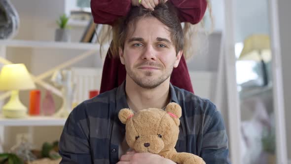 Portrait of Funny Young Man Looking at Camera Holding Teddy Bear As Little Girl Making Ponytail on