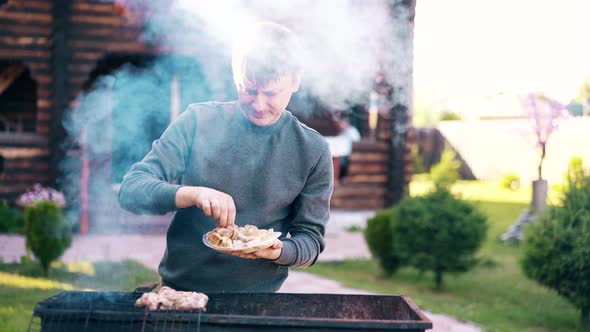 Young Man Stands on the Background of a Summer House Lays Meat on Barbecue Grill