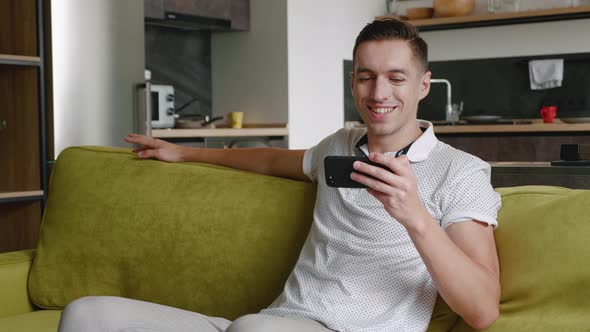 Close Up of Resting Young Man Sitting on the Sofa Watching Video on Smartphone in the Modern