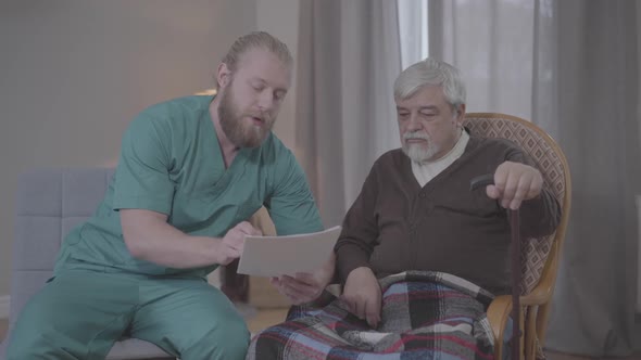 Portrait of Serious Male Nurse in Uniform Reading Documents To Old Caucasian Man Sitting in Armchair