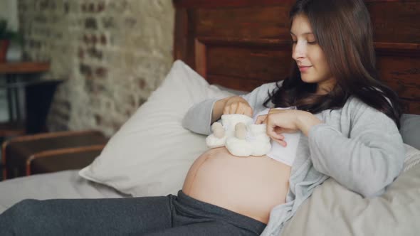 Pregnant Young Lady Is Holding Adorable Baby Shoes in Hands and Making Small Steps on Her Belly
