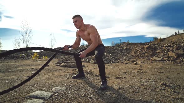 Athletic male performs battle ropes exercise at sunlight. Handsome shirtless man working with battle