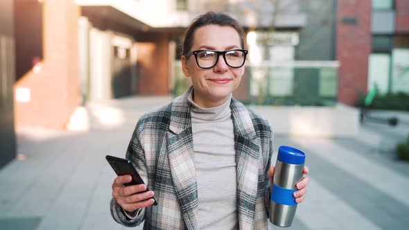 Caucasian Businesswoman with Glasses and a Coat Walks Through the Business District with Thermo Cup