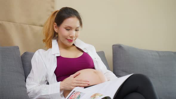 Asian young woman pregnant sitting and reading book with smile on sofa.