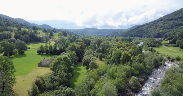 Aerial View Flying Over Meadow and Forest River on Side Mountain in Background Cloudy Sky