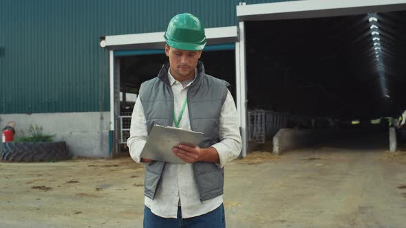 Focused Engineer Making Notes at Modern Cowshed Building in Protective Helmet