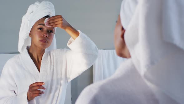 African american woman in bathrobe cleaning her face with cotton pad looking in the mirror at bathro