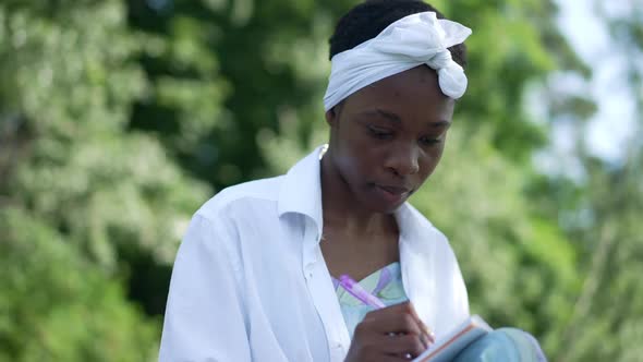 Absorbed African American Young Woman Writing Diary with Pen in Slow Motion Sitting Outdoors