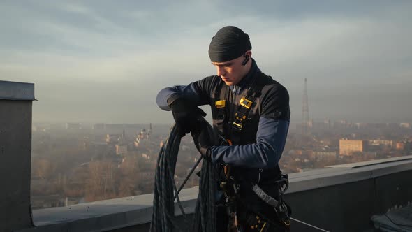 Industrial Professional Male Climber Stands on Rooftop in Uniform and Unravels a Safety Rope for