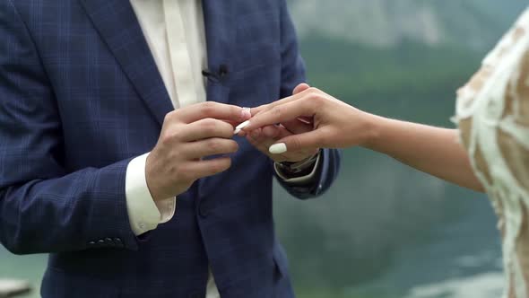 Groom puts bridal ring on bride's finger. Wedding ceremony on lake in mountains, traditional symbols