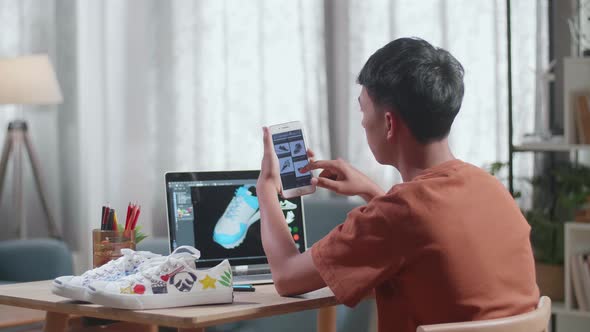 Asian Boy Footwear Designer Looking At Photos On Smartphone While Designing Shoe On A Laptop At Home