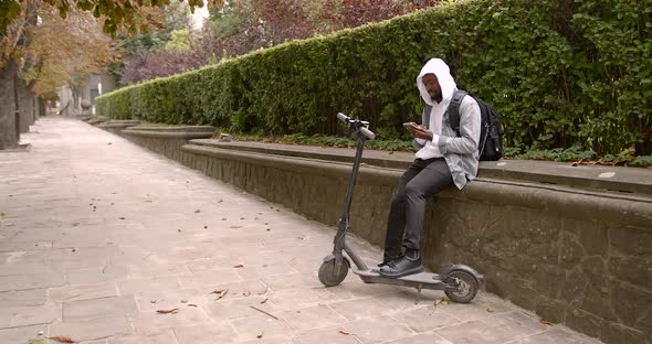 A Young Black Man in a Hood is Talking on the Phone Near an Electric Scooter