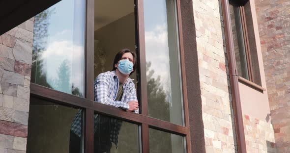 Young Man in Medical Mask Looks Through an Open Window and Talks on Isolation