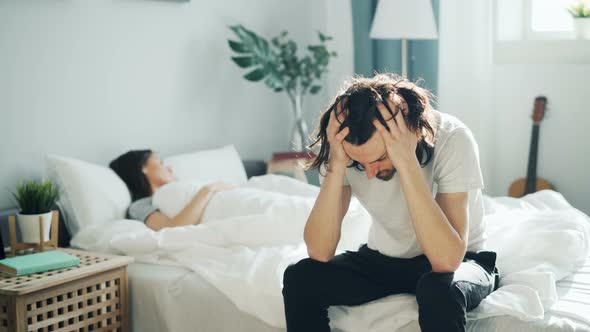 Unhappy Husband Sitting on Bed Touching Sad Face While Wife Sleeping at Home