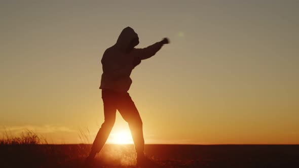 Silhouette of a man in sportswear and a hooded sweatshirt conducts a workout on sunset background