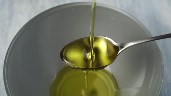 Fresh vegetable oil splashes and overflows a teaspoon and pours into a gray bowl