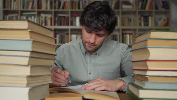 Man Student Sits at a Table in the Library Littered with Books