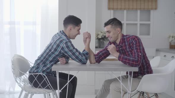 Caucasian Twin Brothers Competing in Armwrestling at Home. Two Positive Adult Brunette Men Sitting