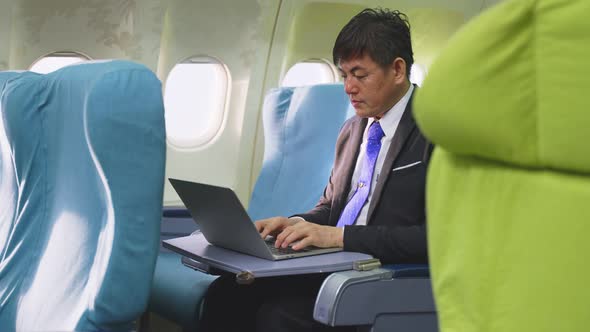 Asian businessman sitting in business class working on laptop on airplane feeling tired on his eyes.