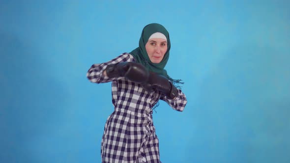 Young Muslim Woman in Boxing Gloves on a Blue Background Looks at the Camera