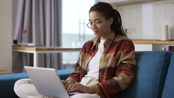 Woman with Dreadlocks is Working on New Project and Using Laptop on Couch in Apartment Room Slow