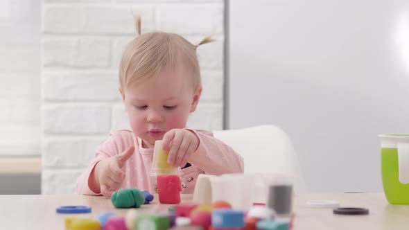 Cute Little Girl with Funny Ponytails on Head Sitting in Kitchen and Playing Multicolored Plasticine