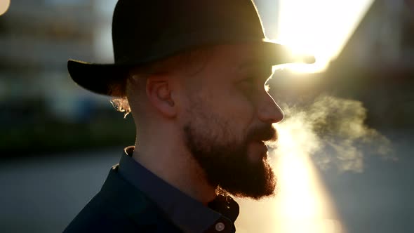 Closeup of the Profile of a Smoking Bearded and Mustachioed Man in a Hat in the Contoured Sunlight