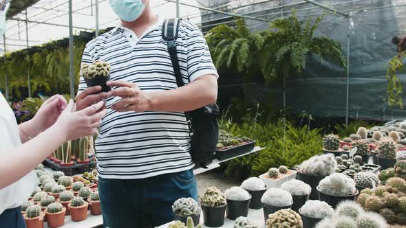 An asian man with a mask on picks up a cactus and gives it to an asian woman, also masked. They are