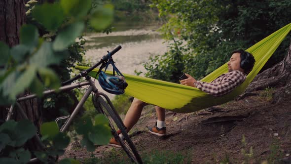 Young Caucasian Man Resting in Hammock Listening to Music on Headphones and Using Smartphone