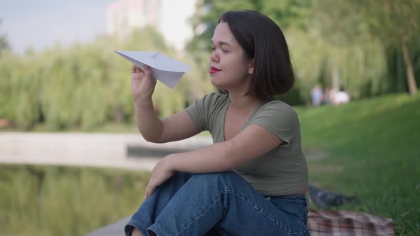 Portrait of Happy Carefree Caucasian Little Woman with Paper Plane Sitting on Lake Shore in Summer