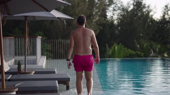 Back View of an Adult Man in Pink Swimwear Trunks Walking By the Lounge Swimming Pool at Sunset in