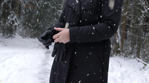 Woman putting on warm gloves on cold winter day in forestry area while snowing