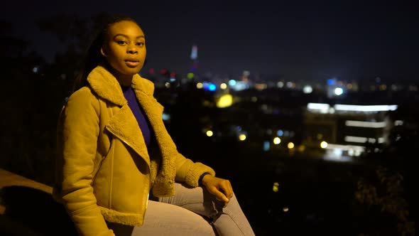 A Young Black Woman Talks to the Camera As She Sits on a Low Wall in an Urban Area at Night