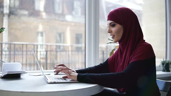 Young Attractive Muslim Girl with Hijab Covering Her Head is Typing Something on Her Laptop While alt