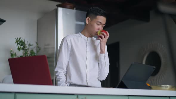 Positive Asian Man Smelling and Biting Healthful Vitamin Apple Smiling Standing in Kitchen in Home