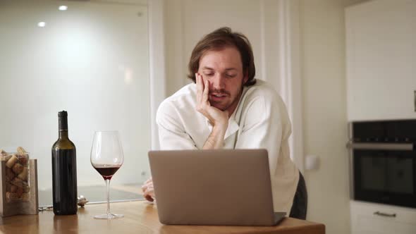 Pensive long-haired man talking by video call on laptop with glass of wine