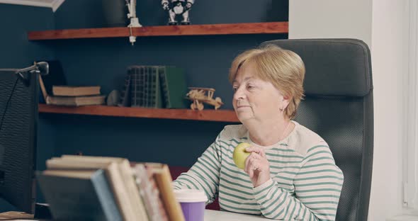 An Elderly Woman Works at Home with a Computer and is About to Eat an Apple
