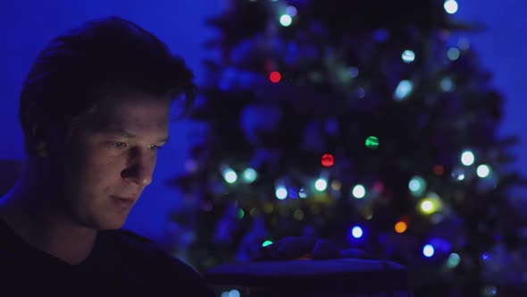 A Man in the Dark Against a Background of Lights Opens a Gift