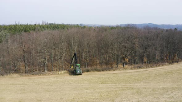Harvester cutting down trees on edge of leafless forest, drone shot.