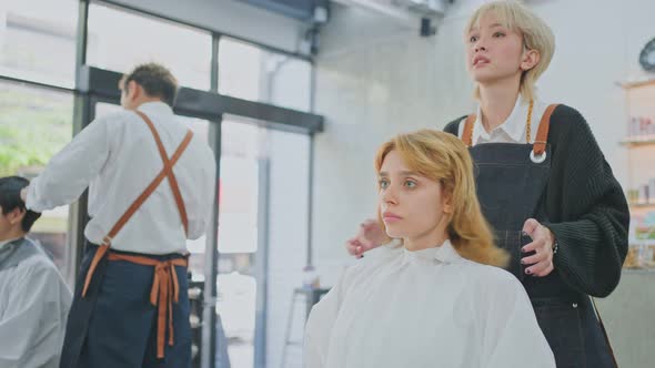 Asian professional hair stylist girl puts on protective cape apron to Caucasian customer in salon.