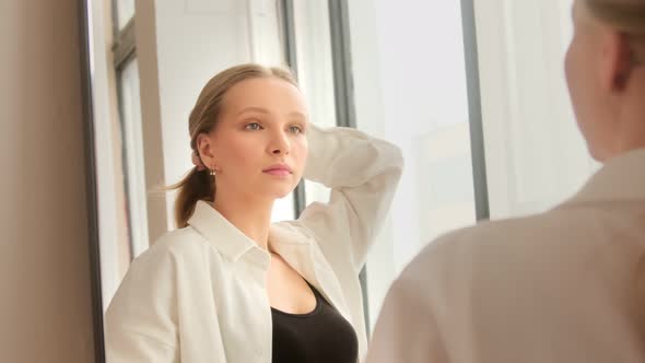 A young blonde woman looking in mirror doing long hair style and getting ready