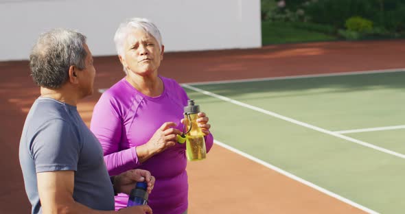 Video of happy biracial senior couple drinking water and talking after training on tennis court