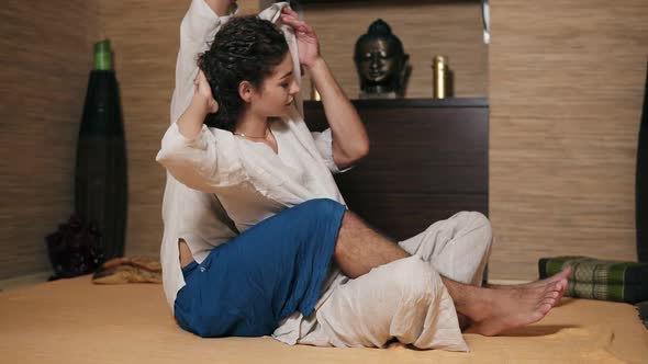 Side View Male Thai Massagist is Stretching Young Woman's Side Sitting Behind Her and Holding Her