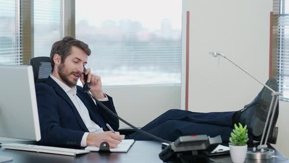 Young Businessman Sitting at the Workplace in Office and Talking on the Phone with His Legs on the