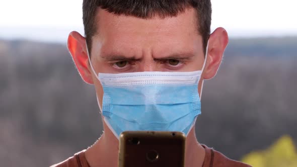 Man in Protective Face Mask Looks at Smartphone Holding It in Hands on Street