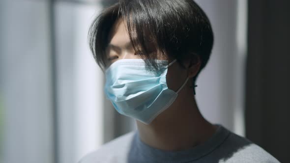 Closeup Face of Young Asian Man in Coronavirus Face Mask Looking Out the Window in Home Office