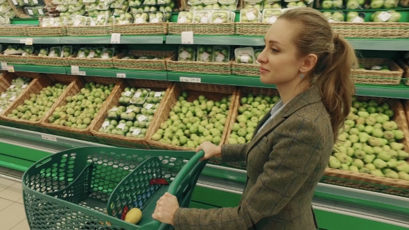 Young Blond Woman Walk Along Shelves With Fruits In Supermarket