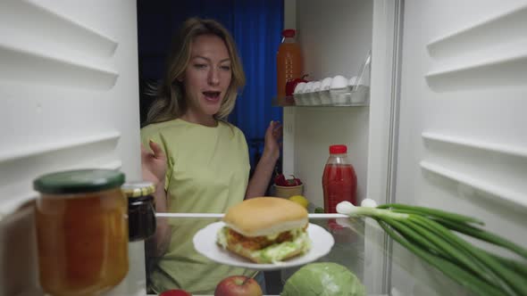 Young Woman Opens the Refrigerator Door at Night and Happy Takes a Burger
