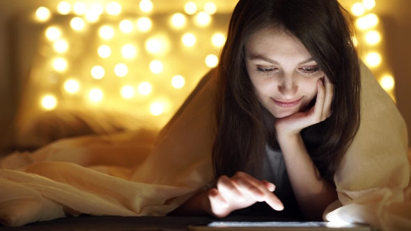 Girl Using Digital Tablet Under Blanket While Lying On Bed At Home